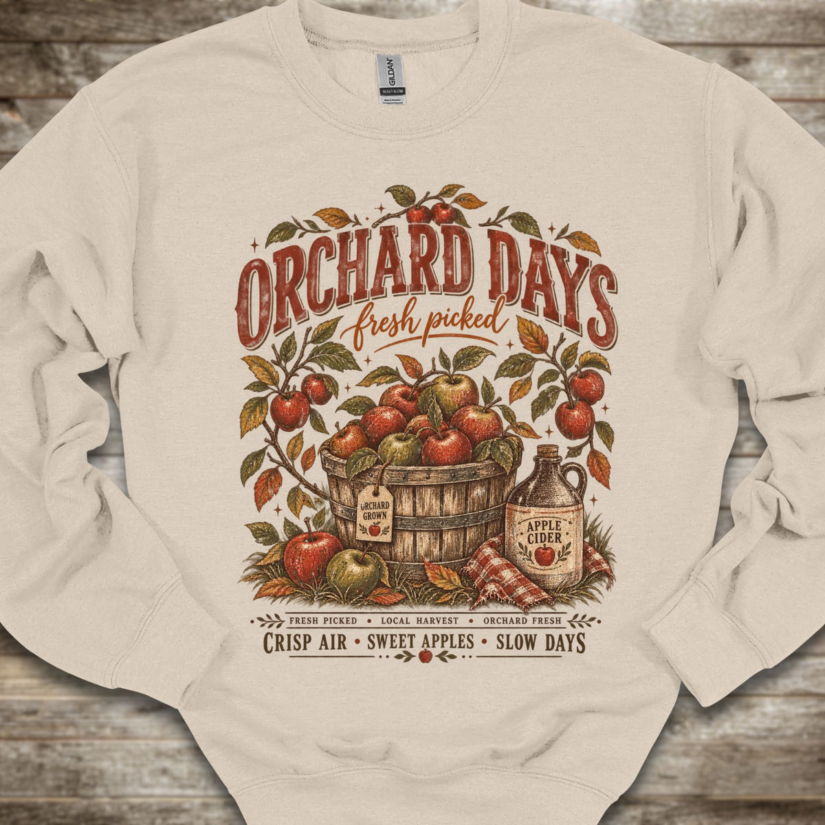 An off-white crewneck sweatshirt with a detailed vintage-style graphic on the front reading 'ORCHARD DAYS fresh picked' above an illustration of apples in a barrel, an apple cider bottle, and text at the bottom, laid flat on a rustic wood background.