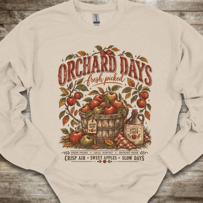 An off-white crewneck sweatshirt with a detailed vintage-style graphic on the front reading 'ORCHARD DAYS fresh picked' above an illustration of apples in a barrel, an apple cider bottle, and text at the bottom, laid flat on a rustic wood background.