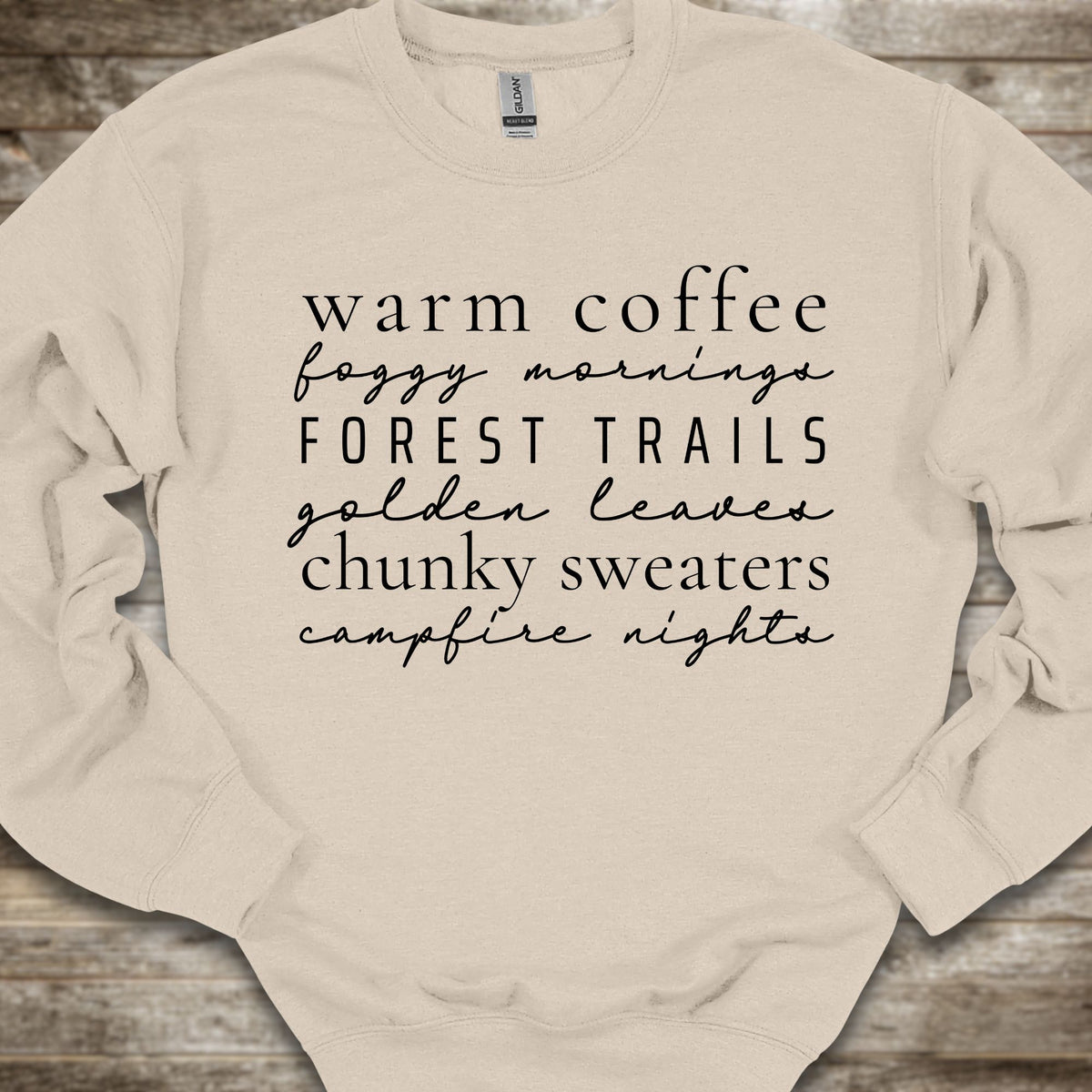 A cream-colored crewneck sweatshirt with black text listing cozy phrases like 'warm coffee', 'foggy mornings', 'FOREST TRAILS', 'golden leaves', 'chunky sweaters', and 'campfire nights', laid flat on a rustic wooden background.