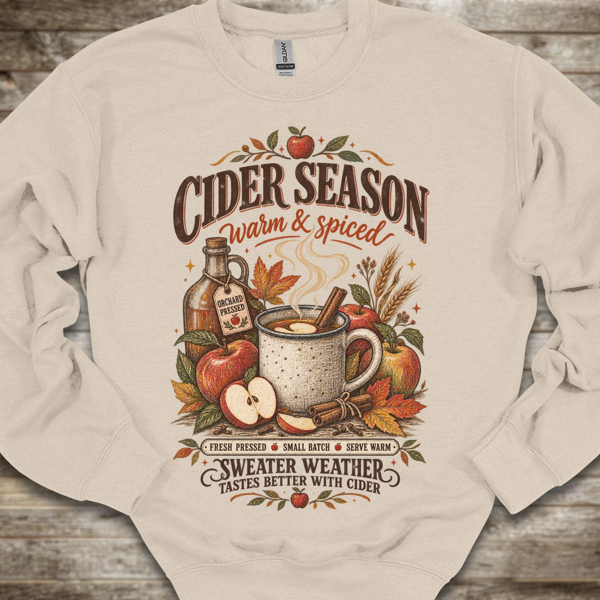 A cream-colored crewneck sweatshirt with a detailed autumn-themed graphic on the front. The graphic features the text 'CIDER SEASON warm & spiced' above an illustration of a mug of cider, apples, cinnamon, and a bottle of cider, with 'SWEATER WEATHER TASTES BETTER WITH CIDER' written below.