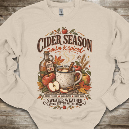 A cream-colored crewneck sweatshirt with a detailed autumn-themed graphic on the front. The graphic features the text 'CIDER SEASON warm & spiced' above an illustration of a mug of cider, apples, cinnamon, and a bottle of cider, with 'SWEATER WEATHER TASTES BETTER WITH CIDER' written below.