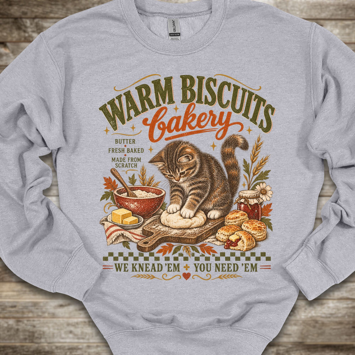 A light heather gray crewneck sweatshirt laid flat on a rustic wooden background, featuring a detailed vintage-style graphic of a tabby cat baking with the text 'WARM BISCUITS bakery' and 'WE KNEAD 'EM + YOU NEED 'EM'.