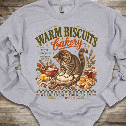 A light heather gray crewneck sweatshirt laid flat on a rustic wooden background, featuring a detailed vintage-style graphic of a tabby cat baking with the text 'WARM BISCUITS bakery' and 'WE KNEAD 'EM + YOU NEED 'EM'.