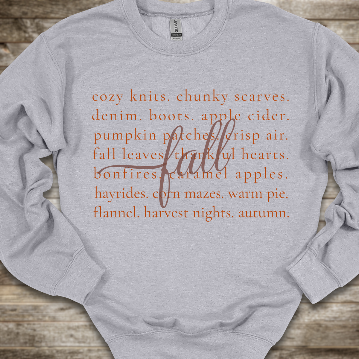 A light heather gray crewneck sweatshirt with orange text listing autumn and cozy items, overlaid with a cursive 'fall' script, laid flat on a rustic wooden background.