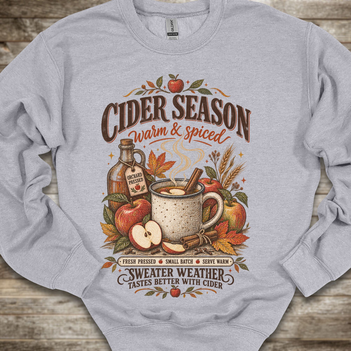 A light gray crewneck sweatshirt laid flat on a rustic wooden surface, featuring a detailed autumn-themed graphic that reads 'CIDER SEASON warm & spiced' and 'SWEATER WEATHER TASTES BETTER WITH CIDER' around an illustration of a mug, cider bottle, apples, cinnamon, and fall leaves.