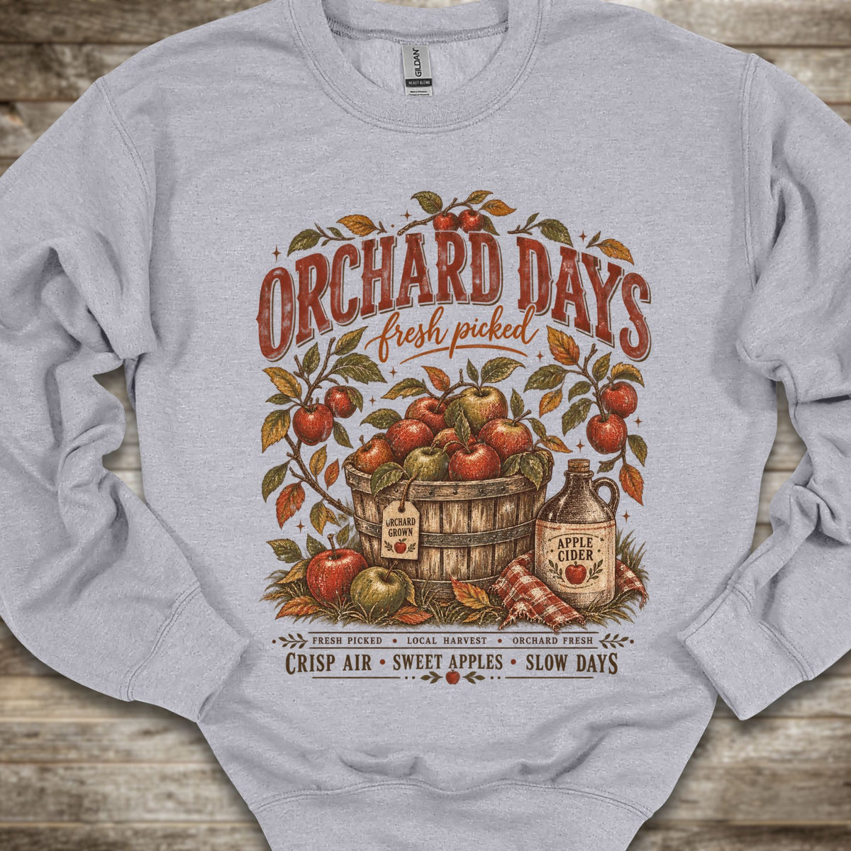 A light gray crewneck sweatshirt with a vintage-style 'Orchard Days' graphic on the front, featuring illustrations of red apples, a wooden basket, an apple cider bottle, and autumn leaves, laid flat on a rustic wood background.