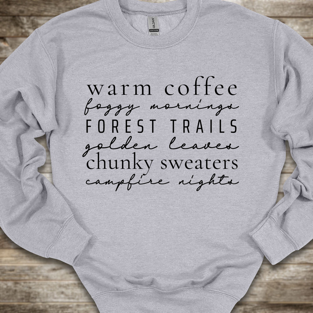 A light heather gray crewneck sweatshirt with black text listing cozy phrases like 'warm coffee', 'foggy mornings', 'FOREST TRAILS', 'golden leaves', 'chunky sweaters', and 'campfire nights', laid flat on a rustic wood background.
