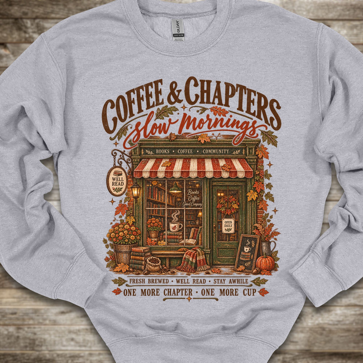 A light gray crewneck sweatshirt featuring a detailed graphic print of a cozy autumn coffee shop with the text 'COFFEE & CHAPTERS Slow Mornings' and additional decorative text, laid flat on a rustic wooden background.