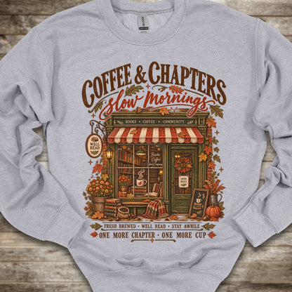 A light gray crewneck sweatshirt featuring a detailed graphic print of a cozy autumn coffee shop with the text 'COFFEE & CHAPTERS Slow Mornings' and additional decorative text, laid flat on a rustic wooden background.