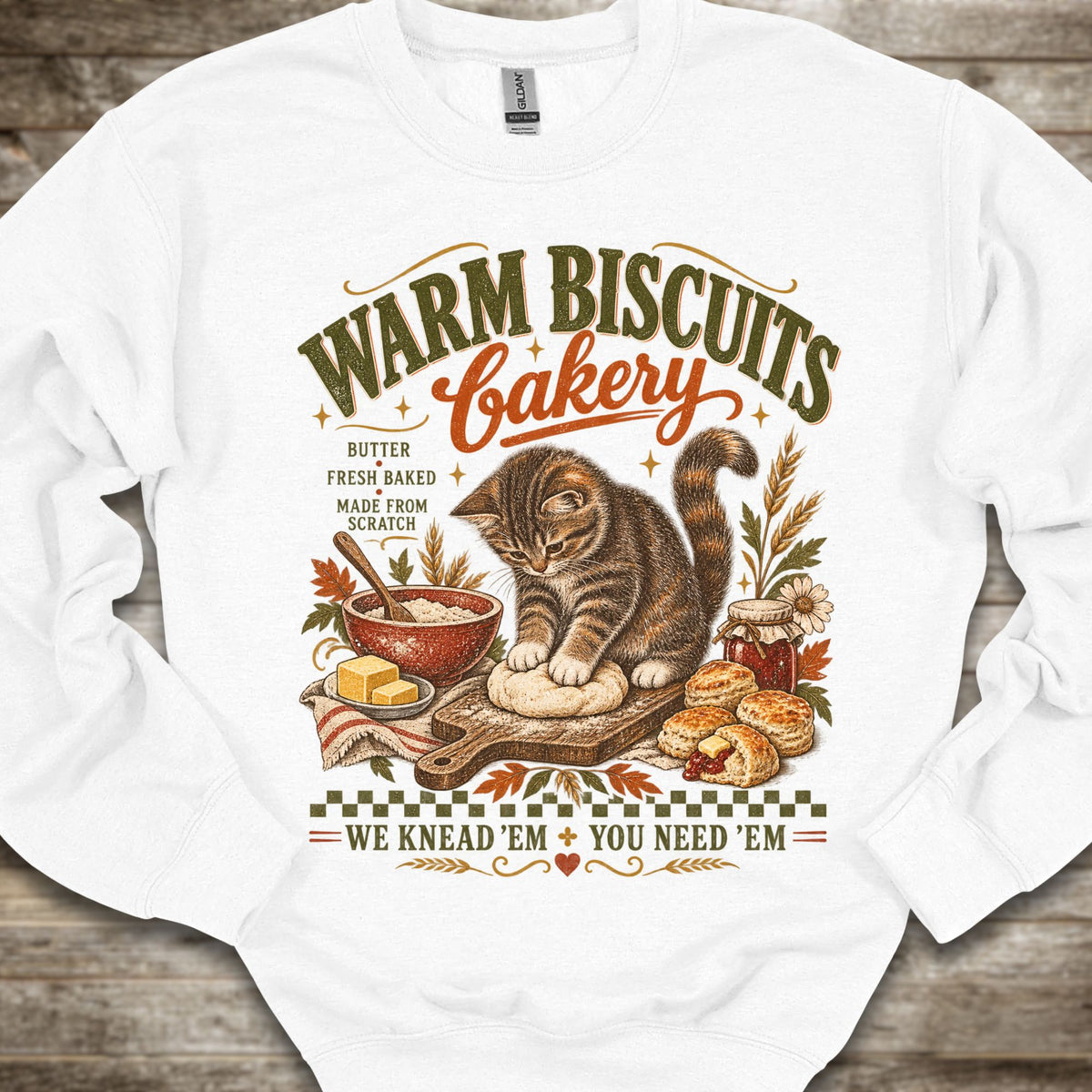 A white crewneck sweatshirt laid flat on a rustic wooden background, featuring a detailed vintage-style graphic of a tabby cat bakery scene with the text 'WARM BISCUITS Bakery' and 'WE KNEAD-EM YOU NEED-EM'.