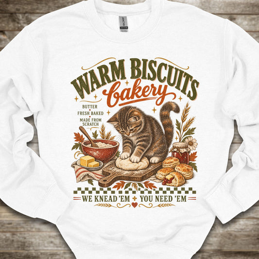 A white crewneck sweatshirt laid flat on a rustic wooden background, featuring a detailed vintage-style graphic of a tabby cat bakery scene with the text 'WARM BISCUITS Bakery' and 'WE KNEAD-EM YOU NEED-EM'.