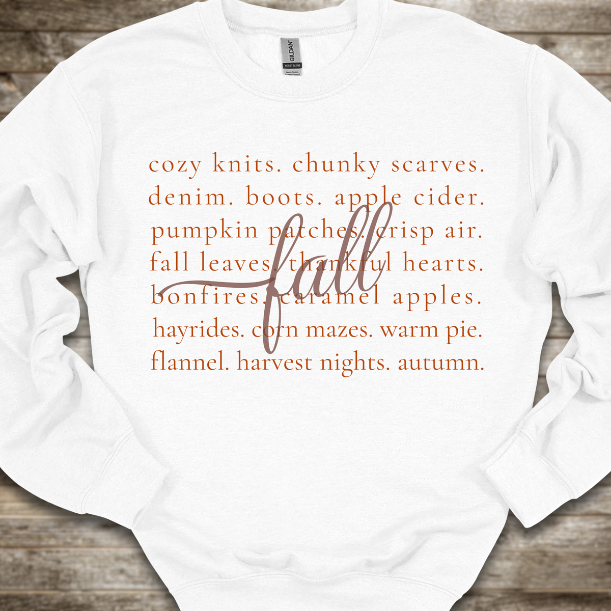 A white crewneck sweatshirt with orange text listing autumn and fall-themed cozy items, overlaid with a large cursive 'fall' script, laid flat on a rustic wooden background.