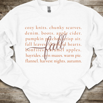 A white crewneck sweatshirt with orange text listing autumn and fall-themed cozy items, overlaid with a large cursive 'fall' script, laid flat on a rustic wooden background.