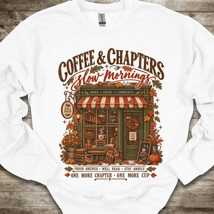 A white crewneck sweatshirt laid flat on a rustic wooden background, featuring a large vintage-style graphic of a cozy coffee shop storefront with the text 'COFFEE & CHAPTERS Slow mornings' and additional autumn-themed decorative text.