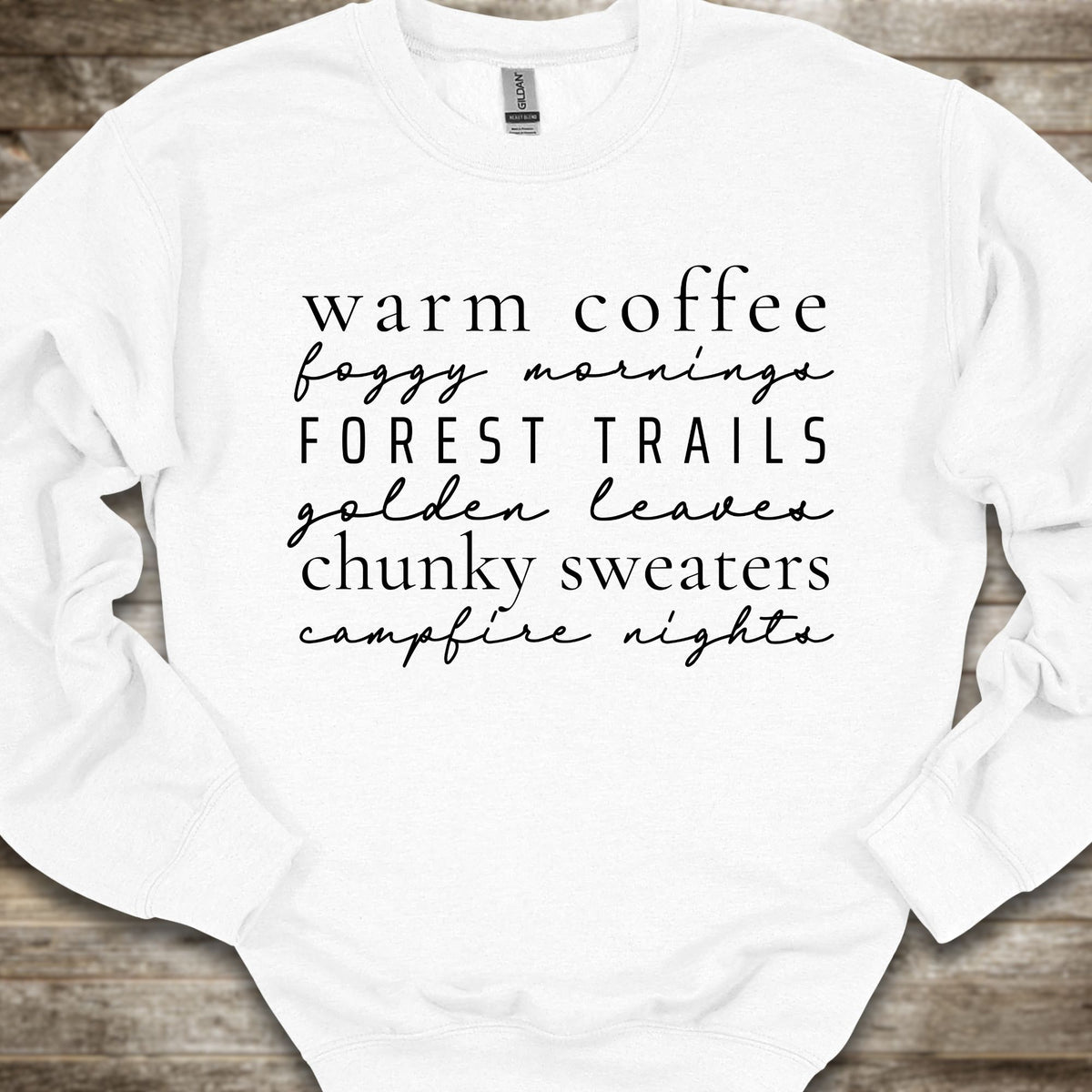 A white crewneck sweatshirt with black printed text listing cozy phrases like 'warm coffee', 'foggy mornings', 'FOREST TRAILS', 'golden leaves', 'chunky sweaters', and 'campfire nights', laid flat on a rustic wood background.