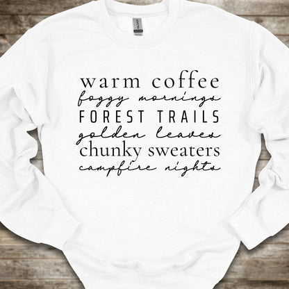 A white crewneck sweatshirt with black printed text listing cozy phrases like 'warm coffee', 'foggy mornings', 'FOREST TRAILS', 'golden leaves', 'chunky sweaters', and 'campfire nights', laid flat on a rustic wood background.