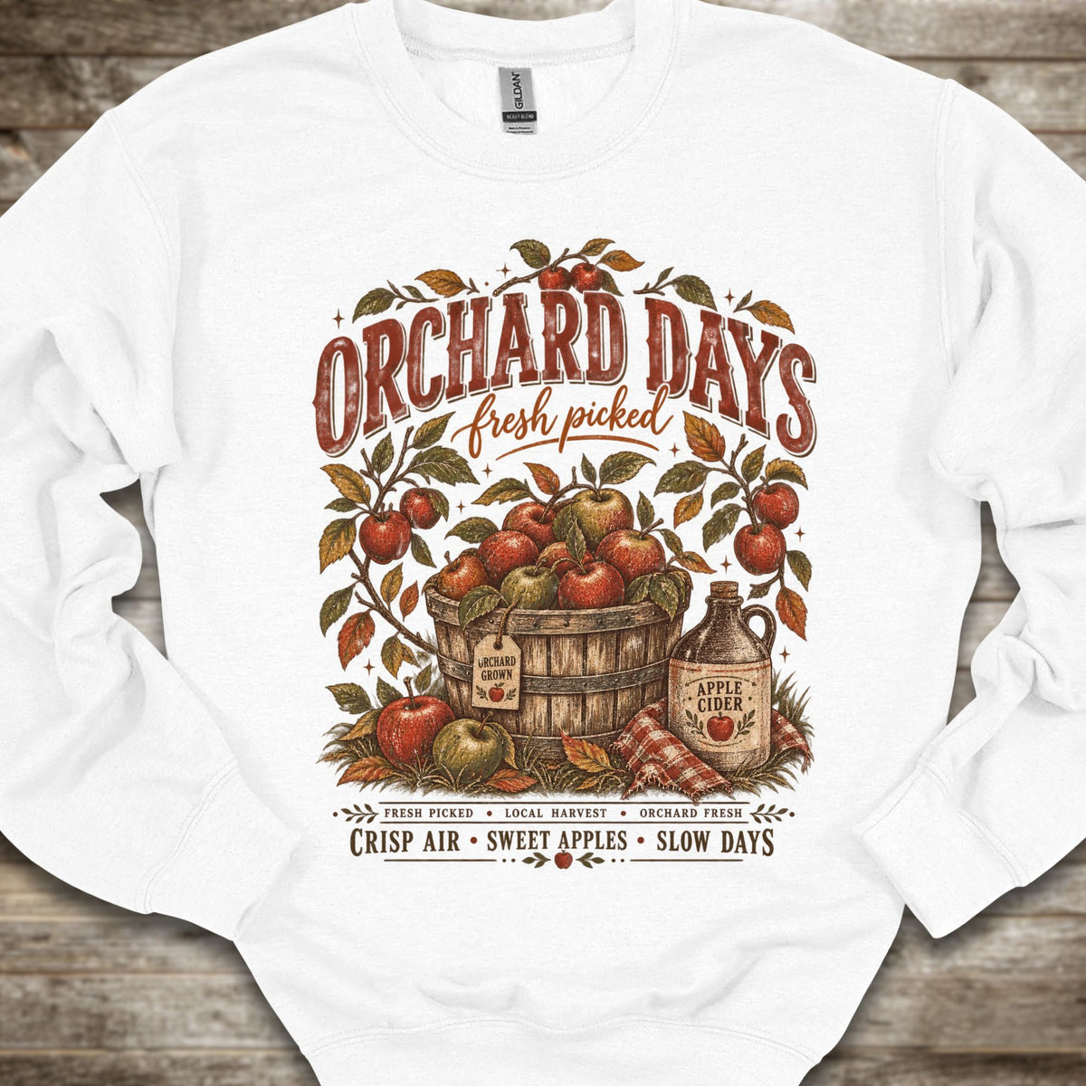 A white crewneck sweatshirt with a vintage-style autumn graphic on the front. The graphic features the text 'ORCHARD DAYS fresh picked' above a wooden barrel filled with red apples, surrounded by leafy branches and an apple cider jug, set against a rustic wood plank background.