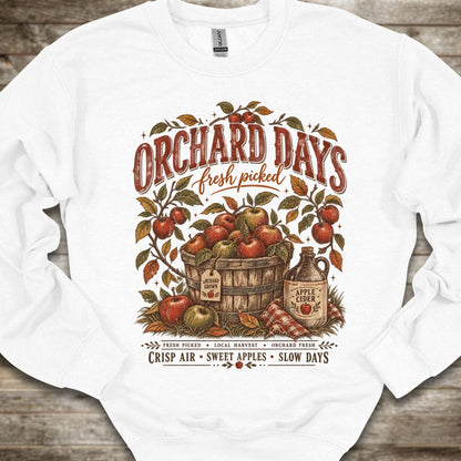 A white crewneck sweatshirt with a vintage-style autumn graphic on the front. The graphic features the text 'ORCHARD DAYS fresh picked' above a wooden barrel filled with red apples, surrounded by leafy branches and an apple cider jug, set against a rustic wood plank background.