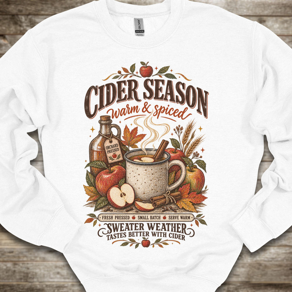 A white crewneck sweatshirt with a detailed autumn-themed graphic on the front. The graphic features the text 'CIDER SEASON warm & spiced' and 'SWEATER WEATHER TASTES BETTER WITH CIDER' surrounding an illustration of a mug of cider, apples, cinnamon, and a bottle of cider.