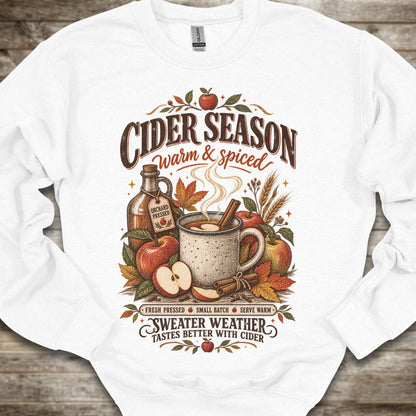A white crewneck sweatshirt with a detailed autumn-themed graphic on the front. The graphic features the text 'CIDER SEASON warm & spiced' and 'SWEATER WEATHER TASTES BETTER WITH CIDER' surrounding an illustration of a mug of cider, apples, cinnamon, and a bottle of cider.