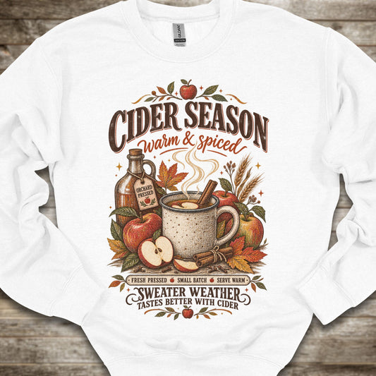 A white crewneck sweatshirt with a detailed autumn-themed graphic on the front. The graphic features the text 'CIDER SEASON warm & spiced' and 'SWEATER WEATHER TASTES BETTER WITH CIDER' surrounding an illustration of a mug of cider, apples, cinnamon, and a bottle of cider.