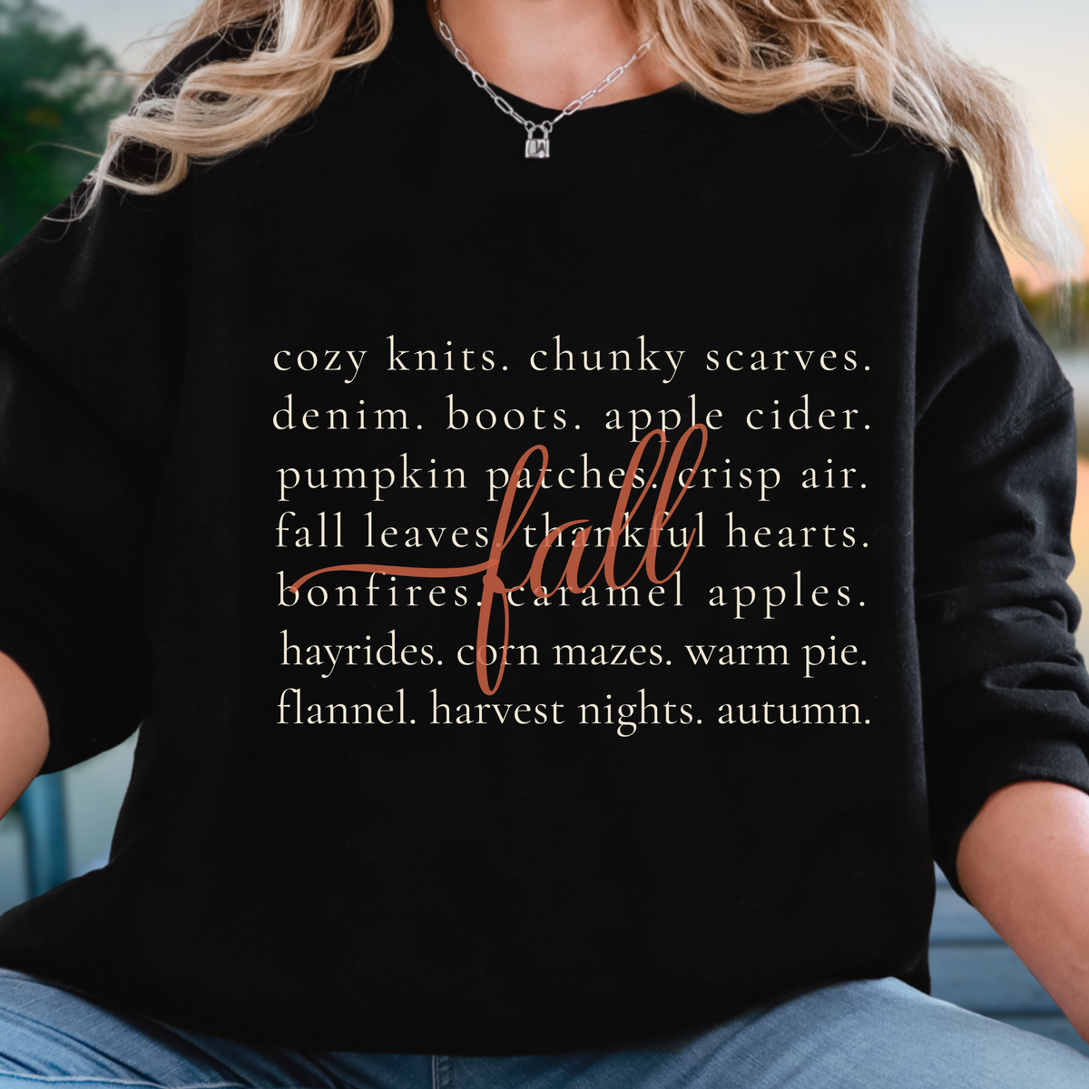 A woman wearing a black long-sleeve t-shirt with a white text graphic listing autumn and fall-themed items, overlaid with a brown cursive 'fall' script. The background is a soft, blurred outdoor setting.