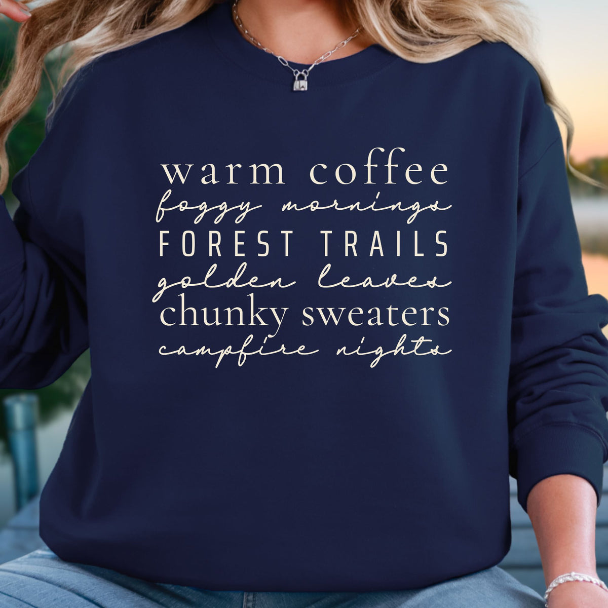 A woman wearing a navy blue crewneck sweatshirt with white text listing cozy phrases like 'warm coffee', 'foggy morning', 'FOREST TRAILS', 'golden leaves', 'chunky sweaters', and 'campfire nights'.