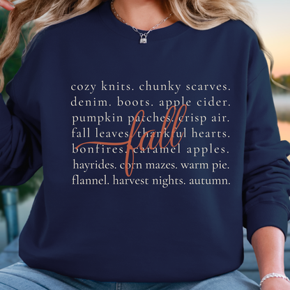 A woman wearing a navy blue long-sleeve sweatshirt with a white text graphic listing autumn themes and a cursive 'fall' word. The photo is cropped from the chest down, showing her blonde hair, necklace, and blue jeans against a softly blurred outdoor background.