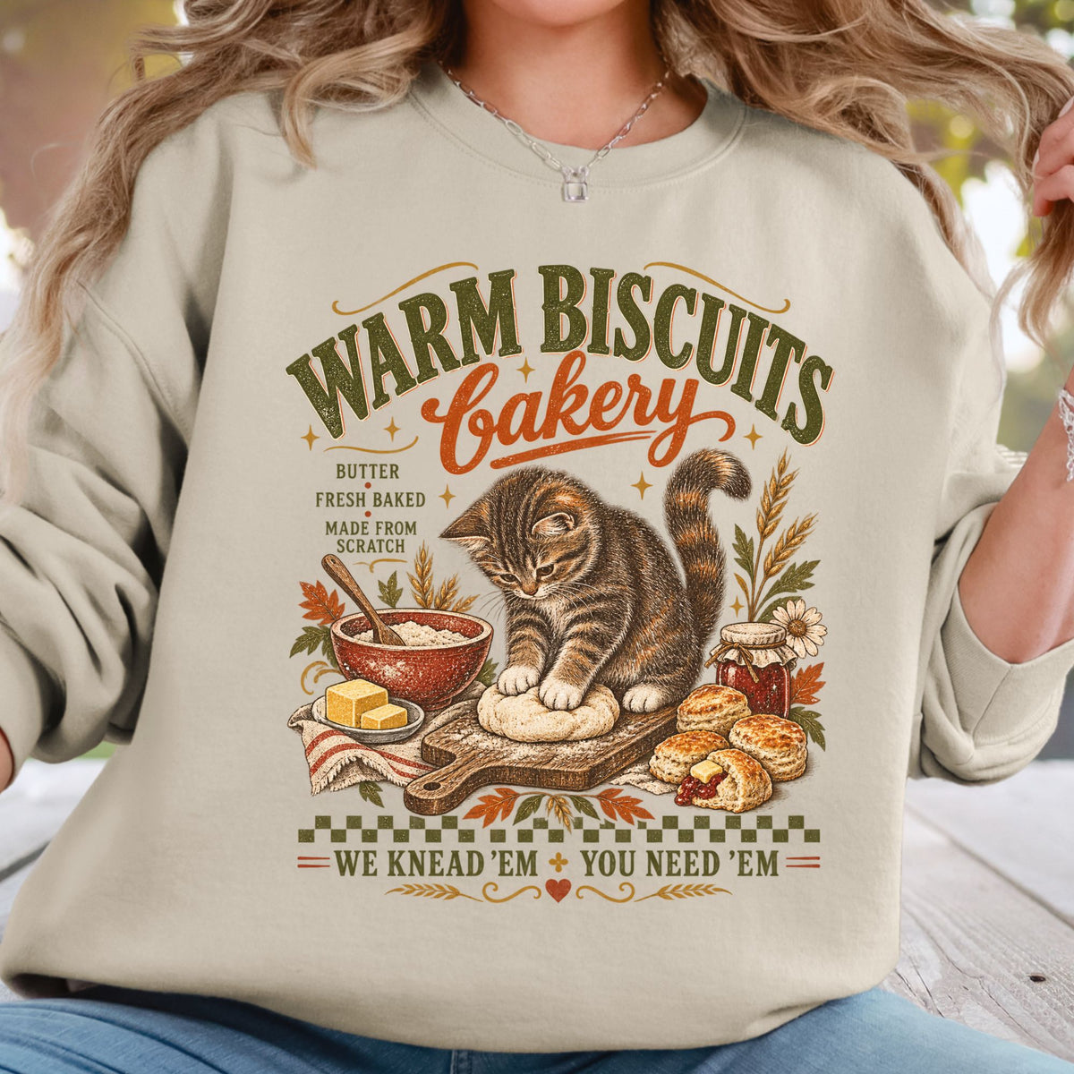 A woman wearing a light beige sweatshirt with a detailed graphic print of a cat baking bread in a bakery scene, with text reading 'WARM BISCUITS Bakery' and 'WE KNEAD 'EM + YOU NEED 'EM'.
