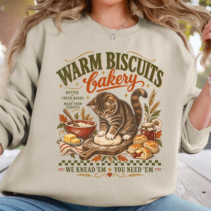 A woman wearing a light beige sweatshirt with a detailed graphic print of a cat baking bread in a bakery scene, with text reading 'WARM BISCUITS Bakery' and 'WE KNEAD 'EM + YOU NEED 'EM'.