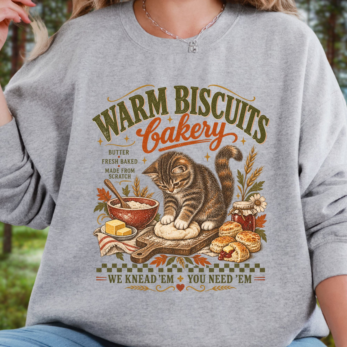 A woman wearing a heather gray long-sleeve sweatshirt with a vintage-style graphic that reads 'Warm Biscuits Bakery' and shows a cat kneading dough with butter, bread, and autumn-themed illustrations.