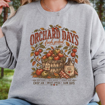 A woman wearing a heather gray long-sleeve sweatshirt with a vintage-style 'Orchard Days' apple graphic on the front, photographed outdoors with blurred trees in the background.