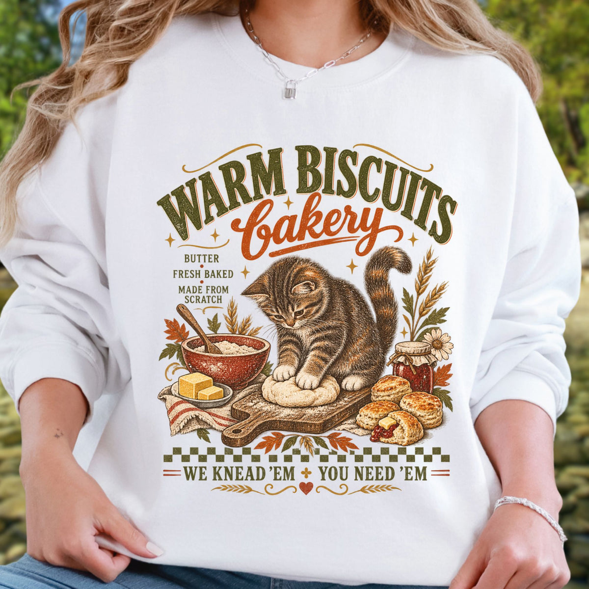 A white crewneck sweatshirt featuring a vintage-style graphic of a cat kneading dough with the text 'WARM BISCUITS Bakery' and 'WE KNEAD 'EM + YOU NEED 'EM', worn by a model in an outdoor setting.