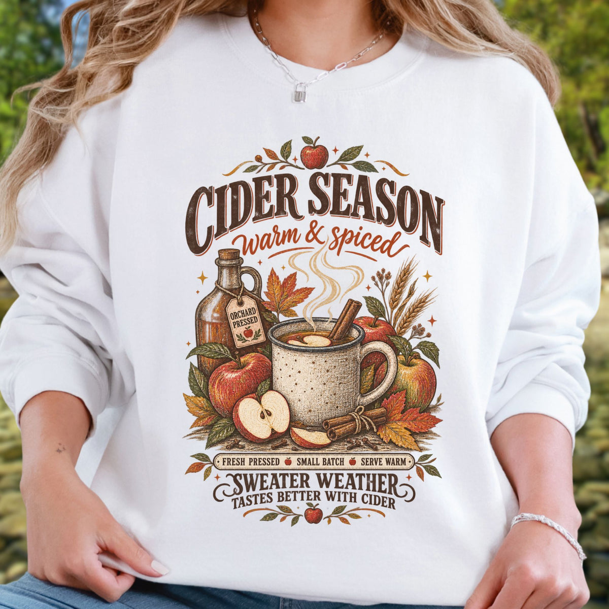 A woman wearing a white long-sleeve crewneck sweatshirt with a detailed autumn-themed graphic that reads 'CIDER SEASON warm & spiced' and 'SWEATER WEATHER TASTES BETTER WITH CIDER.' The print includes an apple, mug, cinnamon stick, bottle, and fall leaves in warm brown, orange, and red tones.