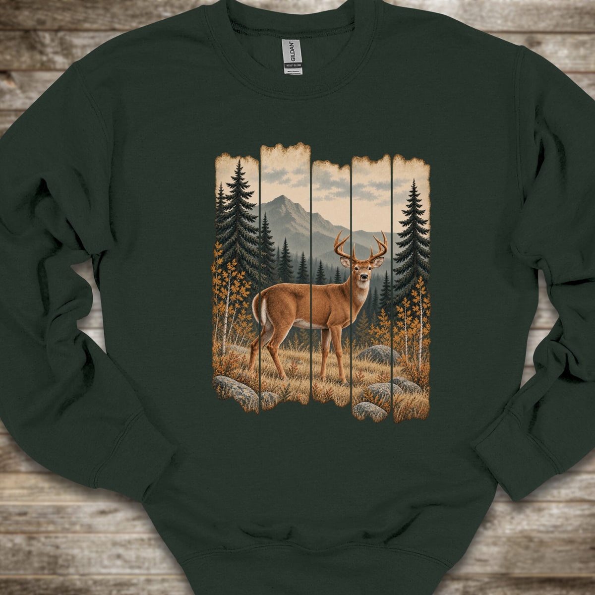 A dark forest green crewneck sweatshirt laid flat on a rustic wooden background, featuring a large graphic print of a deer in a mountain pine forest.