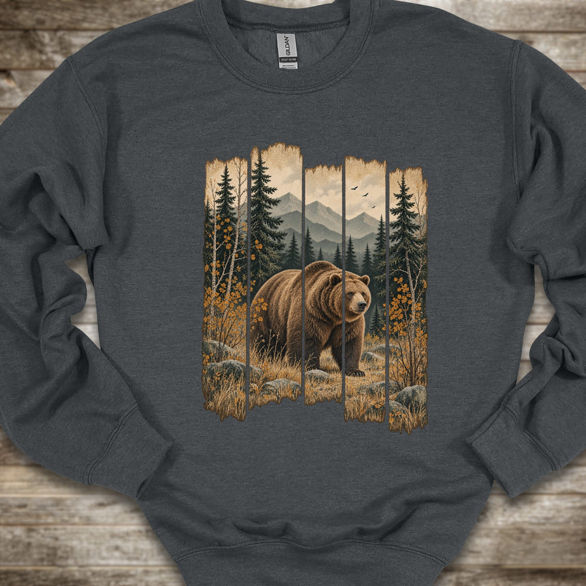 A dark gray crewneck sweatshirt featuring a large graphic print of a brown grizzly bear in an autumn forest with mountains in the background, laid flat on a rustic wooden surface.