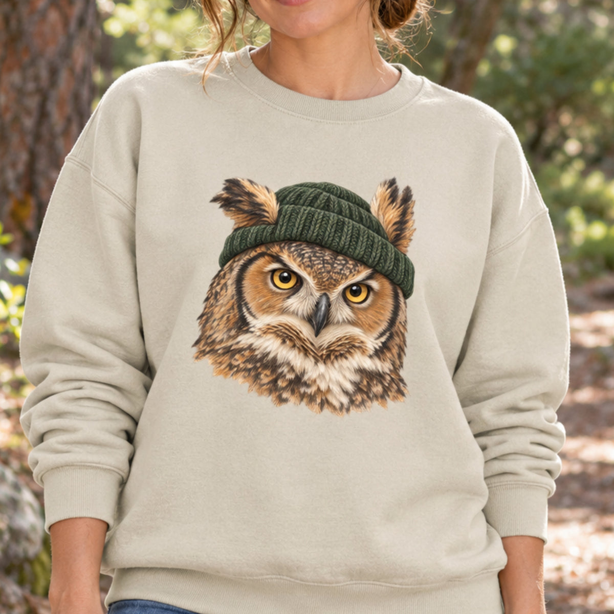 A woman wearing a light beige crewneck sweatshirt with a large graphic of an owl wearing a dark green knit beanie on the front, set against an outdoor blurred background of trees and sunlight.
