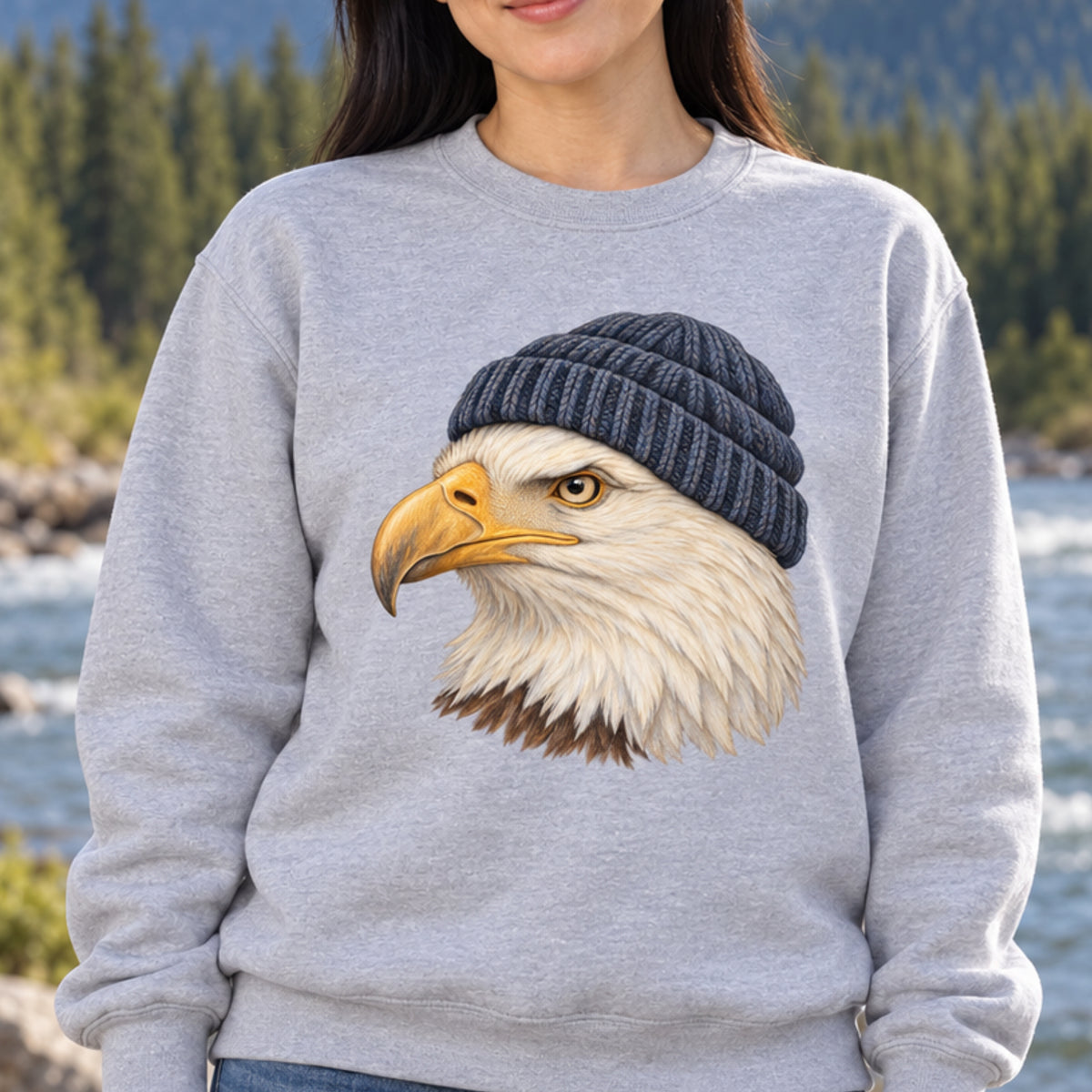A woman wearing a light gray crewneck sweatshirt with a graphic of a bald eagle wearing a dark gray beanie, set against a blurred background of a river and pine trees.
