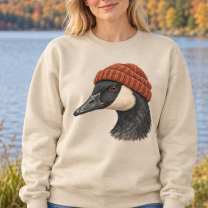 A woman wearing a cream-colored crewneck sweatshirt with a graphic of a Canada goose wearing a rust-orange knit beanie, set against a blurred background of a lake and autumn trees.